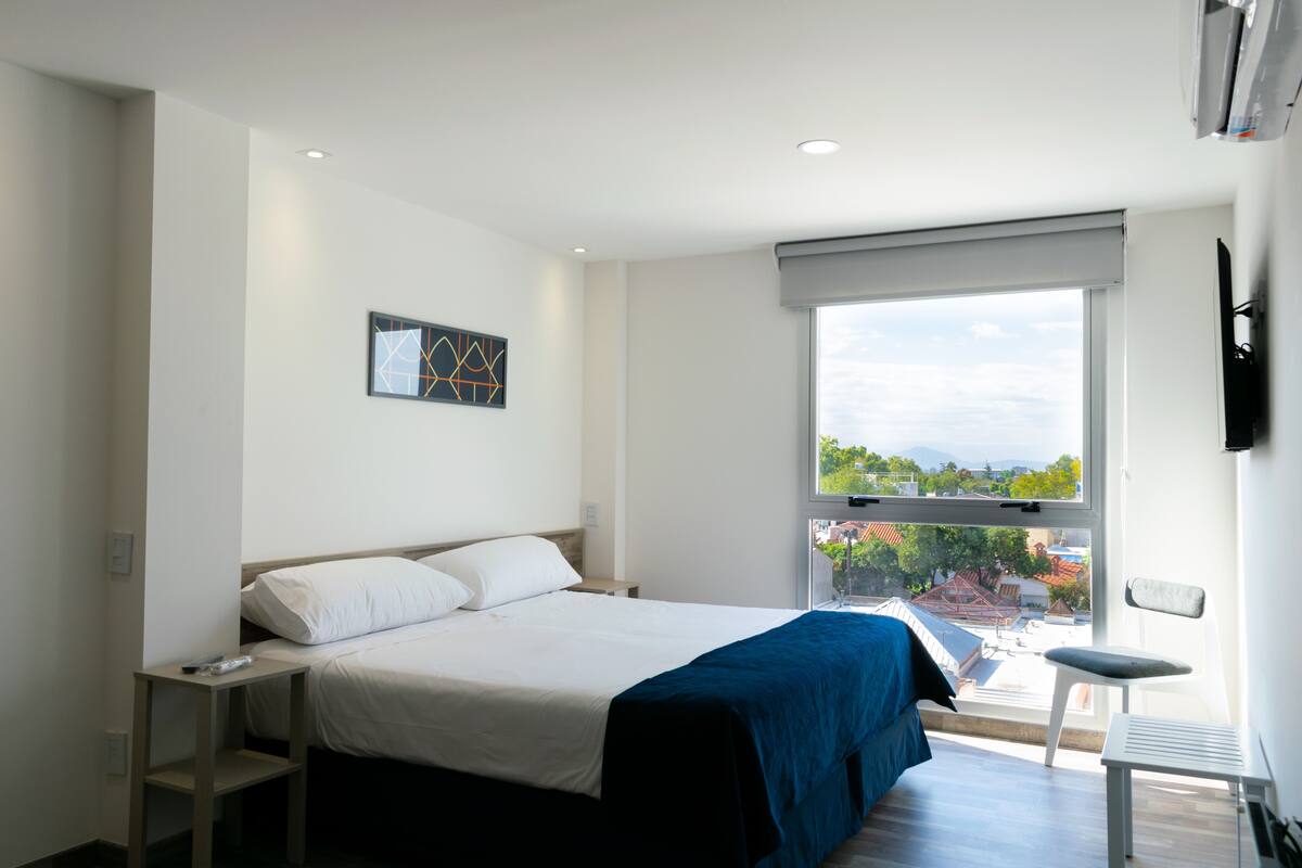 A modern hotel room with a large bed, a small table, a chair, and a window with a city view.