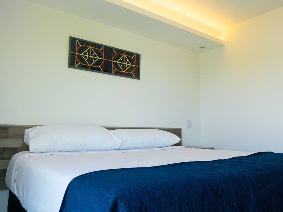 Una cama bien tendida con ropa de cama blanca y azul, un cabecero de madera y una obra de arte geométrica en la pared.