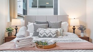 A neatly made bed with pillows, a bedside lamp, a small plant, and a basket of folded towels.