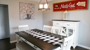 Een eetruimte met een geruit tafelkleed, witte stoelen, een kroonluchter, een vintage bord met '50s North East erop, en een vaas met bloemen aan de muur.