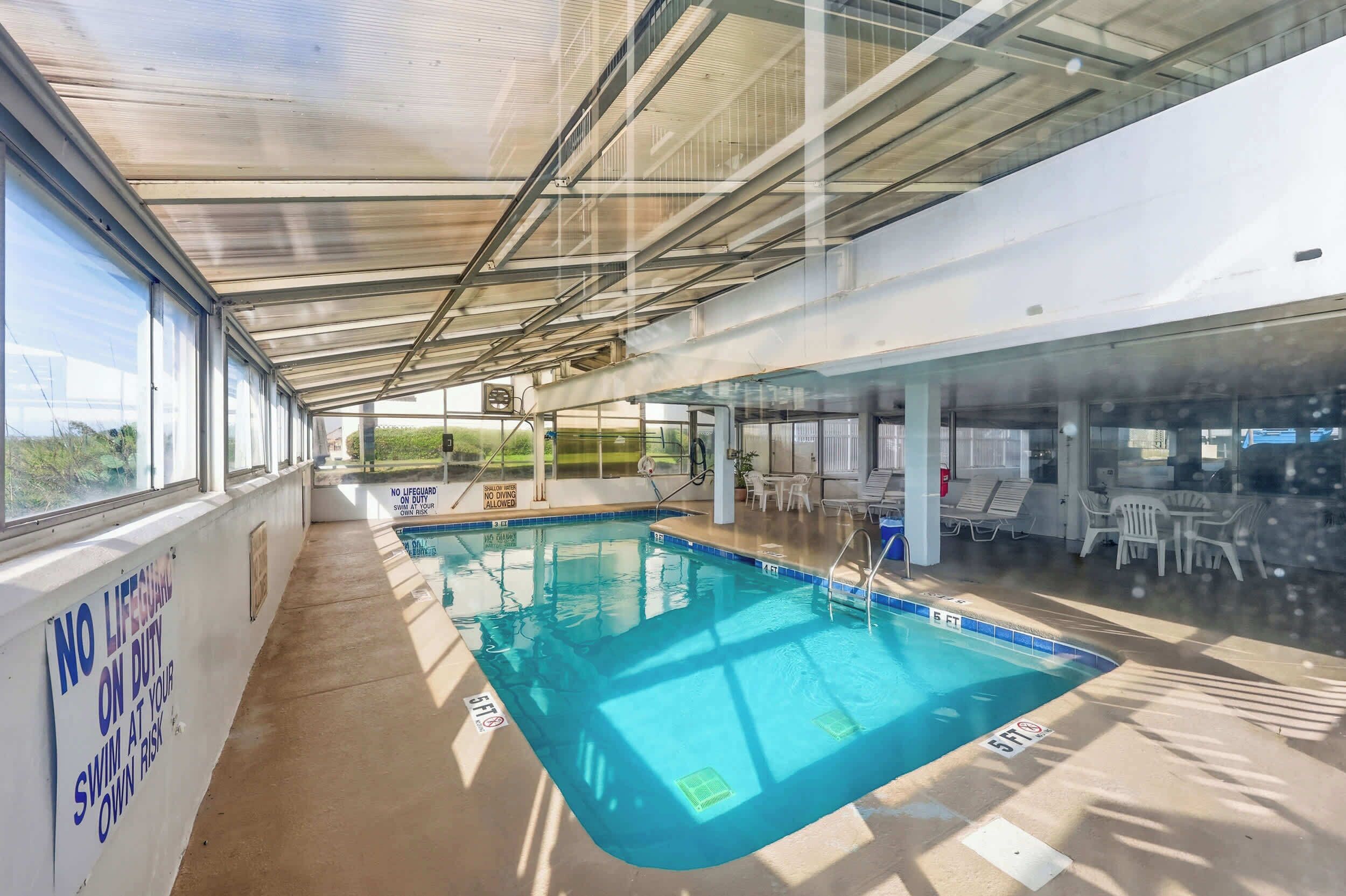 Une piscine intérieure avec des parois en verre transparent, un espace aménagé au bord de la piscine avec des chaises longues, et un panneau d’avertissement indiquant les risques liés à la baignade.