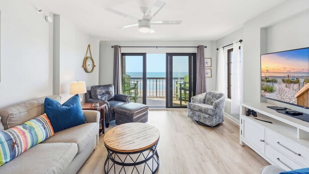 A living room with a sofa, armchair, ottoman, round coffee table, TV, and a balcony with a view. - Sandy Bits Oceanfront Premium Condo with Pool (North Myrtle Beach)