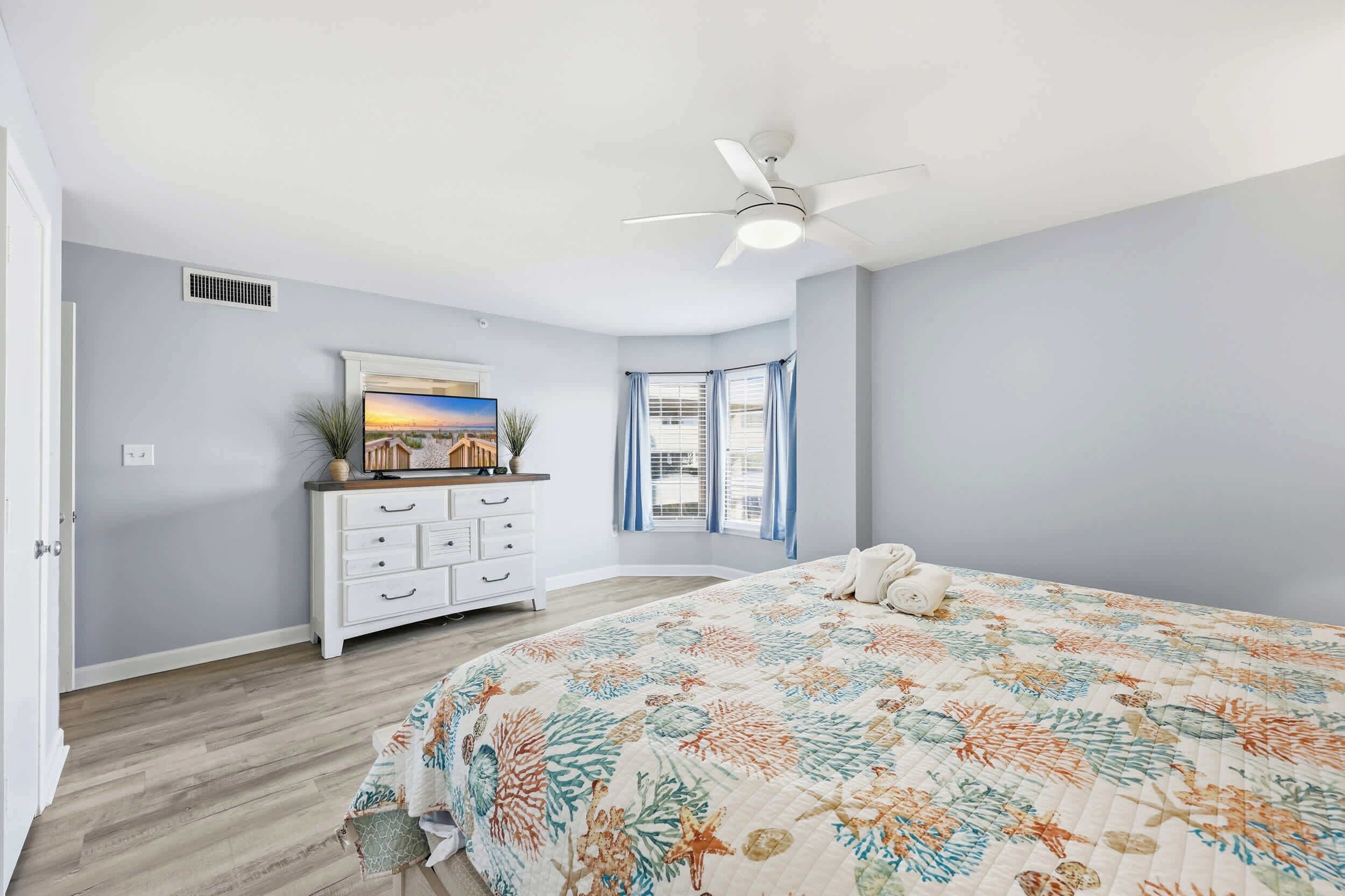 Une chambre à coucher avec un lit, une commode, un ventilateur de plafond, une fenêtre avec des rideaux et un tableau au mur.