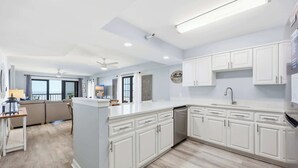 A modern kitchen with white cabinetry, a central island, and a dining area with a sofa. - Sandy Bits Oceanfront Premium Condo with Pool (North Myrtle Beach)