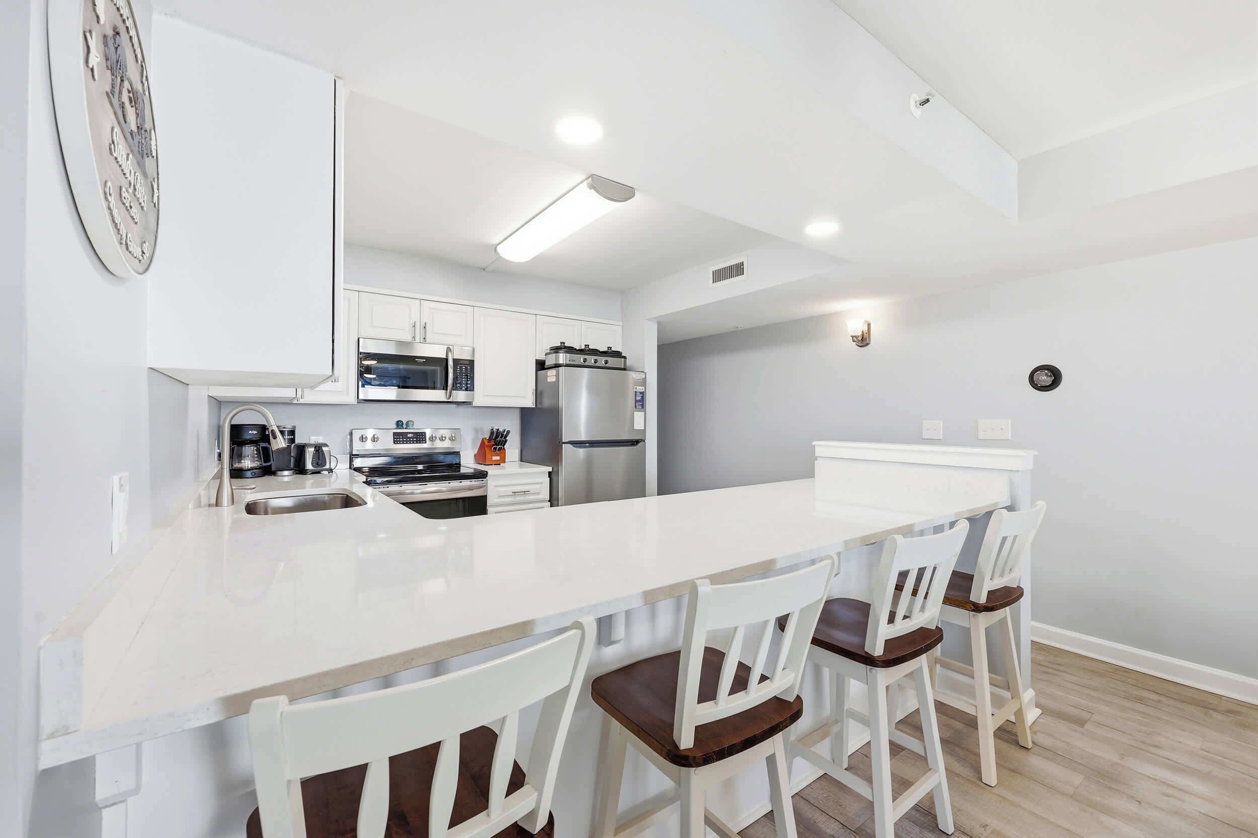 Uma cozinha moderna com armários brancos, uma ilha central com banquetas, micro-ondas, frigorífico e máquina de café.