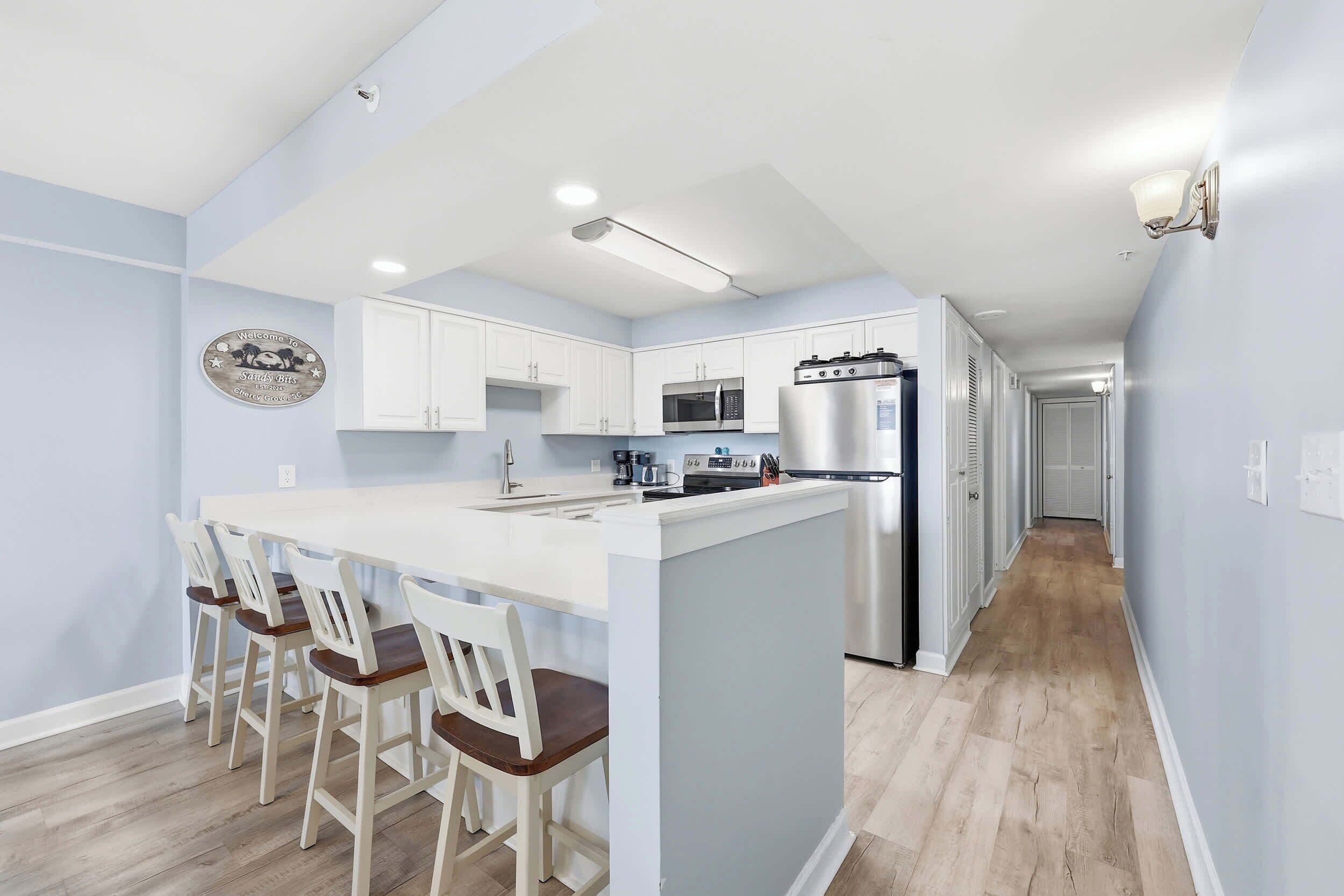 A modern kitchen with white cabinets, a central island, bar stools, a microwave, a refrigerator, and a wall-mounted light fixture.