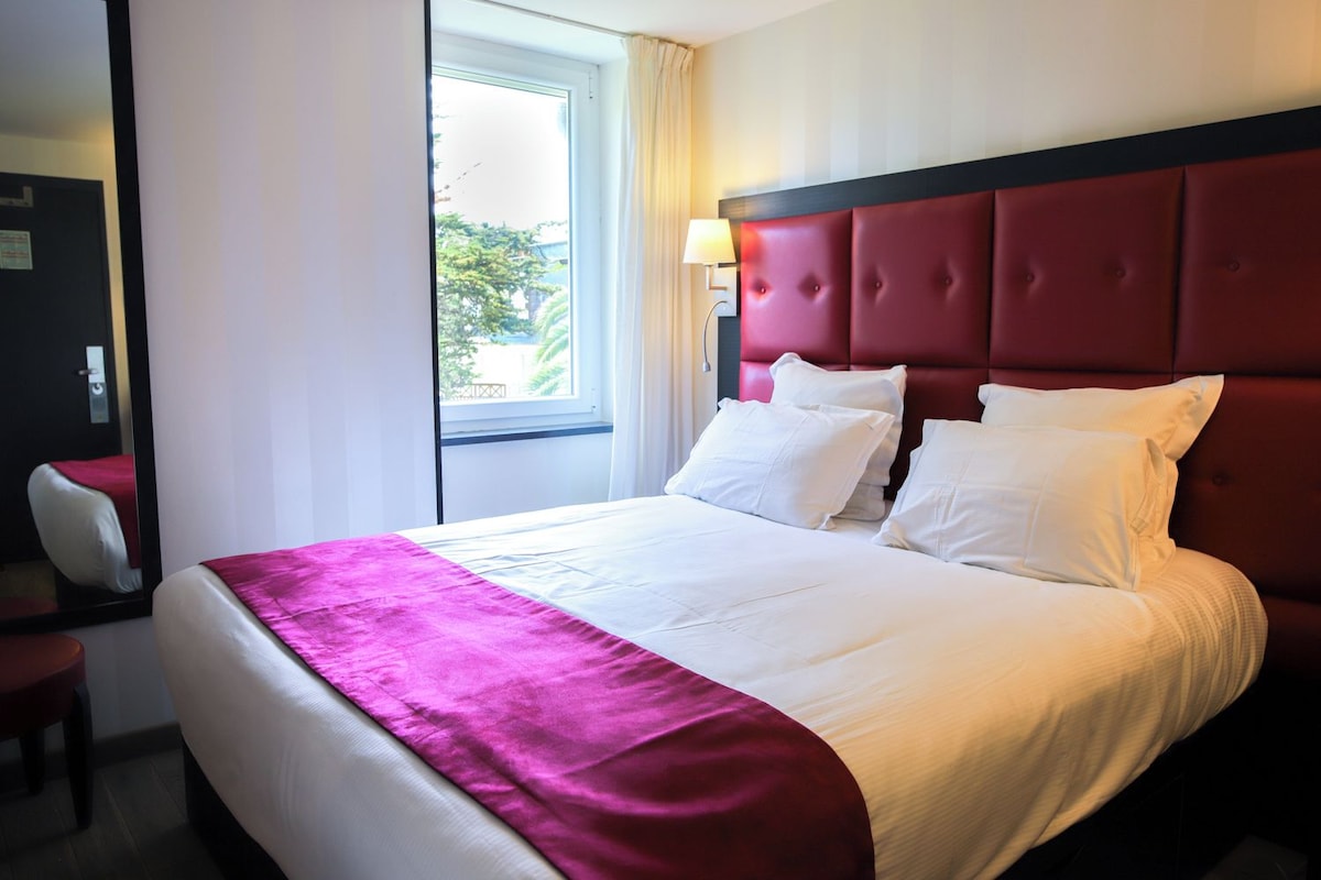 Una habitación de hotel con una cama grande, una ventana con cortinas, una mesita de noche y una silla.