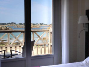A room with a view of a beach and rocky coastline, a wooden deck outside, and a bed with a white pillow. - Logis Hôtel Saint-Guirec et de la Plage (Perros-Guirec)