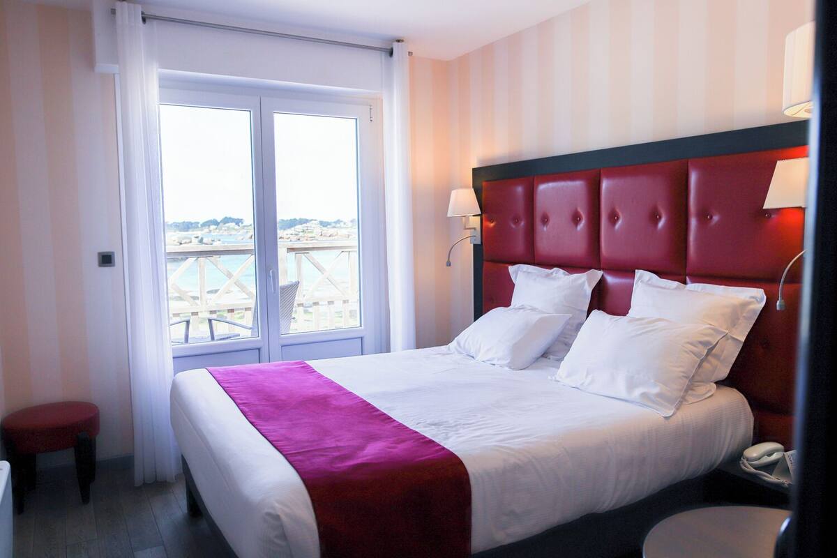 A hotel room with a bed, a bedside table, a lamp, and a view of the sea.