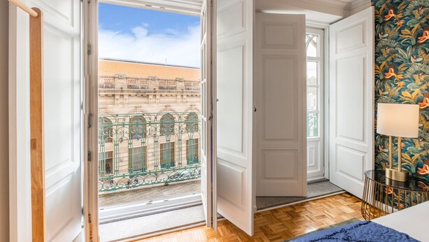 A room with a wooden floor, a bed, a window with a view of a building, and a door leading outside. - São Bento Deluxe 42 (Porto)