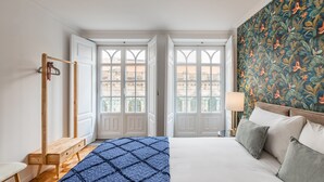 A bedroom with a bed, a wooden bench, a hanging rack, and a window with a view of buildings. - São Bento Deluxe 42 (Porto)