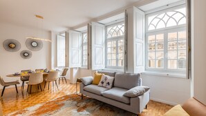 A modern living room with a grey sofa, a dining table with chairs, and large windows with white frames. - São Bento Deluxe 42 (Porto)