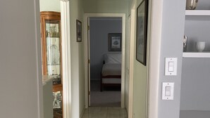 Ein schmaler Flur mit einem Bett am Ende, einer Tür links und einem Regal mit Gegenständen rechts.