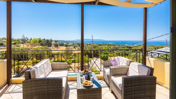 Un balcone con mobili in vimini e un tavolino in vetro, con vista su un paesaggio suggestivo, ricco di vegetazione e affaccio sul mare.