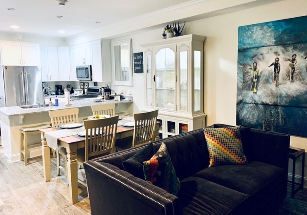 A modern kitchen with white cabinets, a dining area with wooden chairs, and a black sofa with colorful pillows.