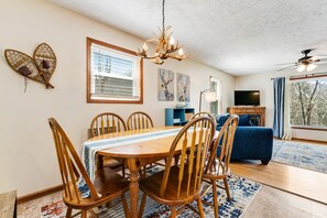 A dining area with a wooden table and chairs, a chandelier, artwork on the walls, and a television in the background.