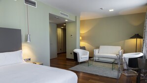 A modern hotel room with a large bed, a white sofa, a clear chair, and a desk.