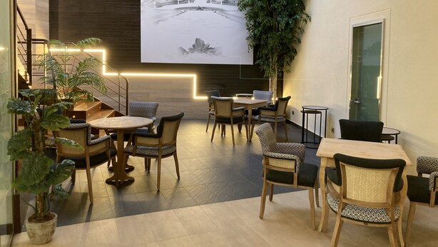 A modern interior with a large screen displaying a building, surrounded by seating areas and potted plants.