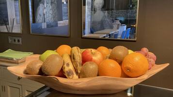 A wooden bowl filled with various fruits on a table in a room with two windows.