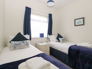 Quarto duplo com duas camas, mesa de cabeceira, luminária e janela com cortinas.