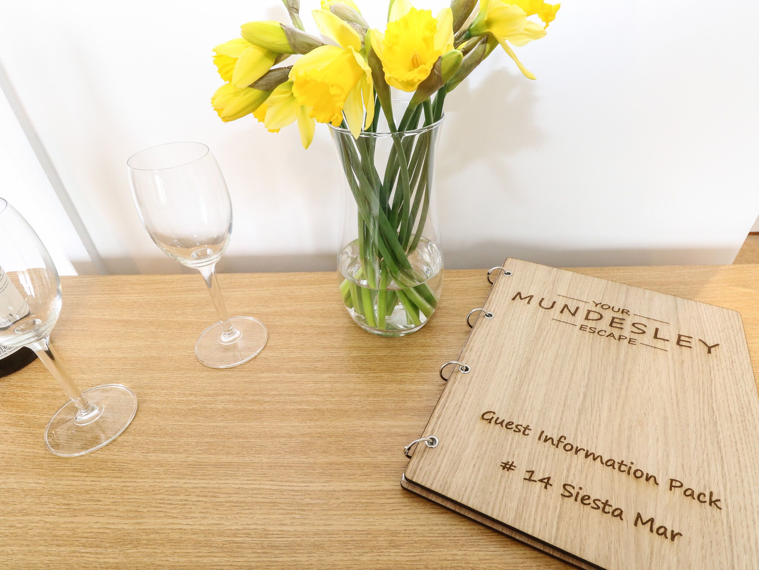 Un présentoir en bois contenant des informations pour les clients, avec un texte gravé, un vase en verre transparent avec des fleurs jaunes et deux verres à vin vides, le tout posé sur une table en bois.