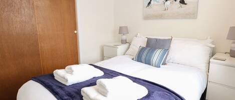 A neatly made bed with white linens and a blue throw blanket, a wooden door, and a wall-mounted painting of birds.