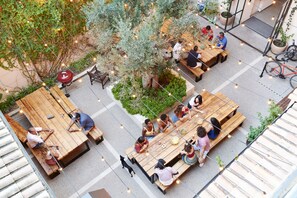 Un área de comedor al aire libre con mesas y bancos de madera, rodeada de follaje e iluminación con luces colgantes.