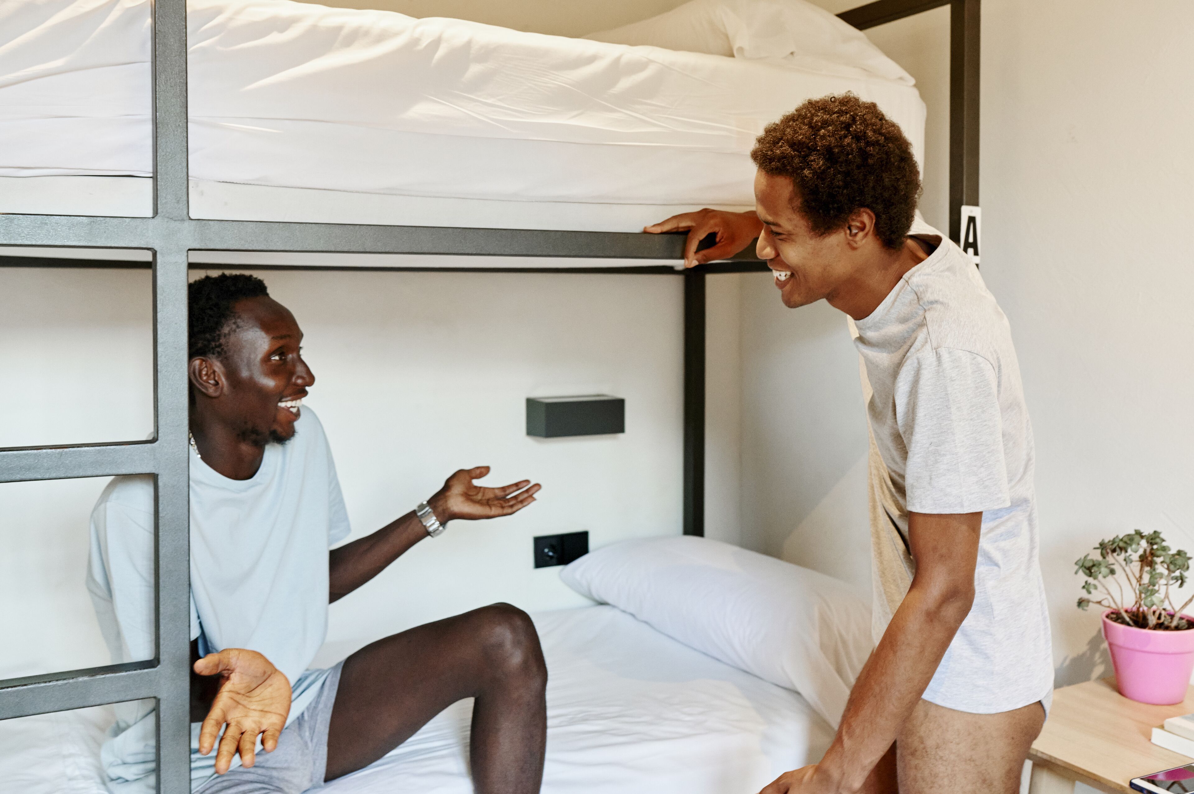 Una cama litera con dos personas, una sentada y otra de pie, en una habitación con una mesita y una planta en maceta.
