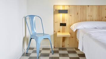 Una habitación con piso a cuadros, una silla azul, una cama con ropa de cama blanca y un cabecero de madera con repisa y lámpara.