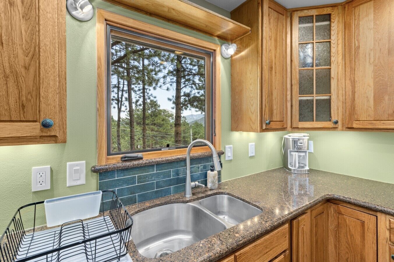 A kitchen with a double sink, wooden cabinets, a window with a view of trees, and a coffee maker on the counter.