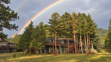 A two-story cabin with a deck, surrounded by tall pine trees, under a rainbow.