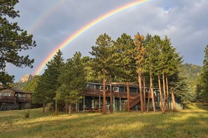 A two-story cabin with a deck, surrounded by tall pine trees, under a rainbow.