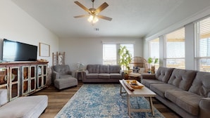 Un salon avec un ventilateur de plafond, un téléviseur, un canapé gris, une table basse et un tapis à motifs.