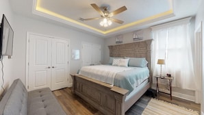 Une chambre à coucher comprenant un lit, un ventilateur de plafond, une table de chevet, une lampe et une fenêtre avec des rideaux.