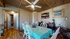 A dining area with a table set for four, white chairs, a ceiling fan, wooden walls, and a kitchen area visible through an open door. - The Mango Hut: 2  BR, 1  BA House in Port Bolivar, Sleeps 8 (Port Bolivar)