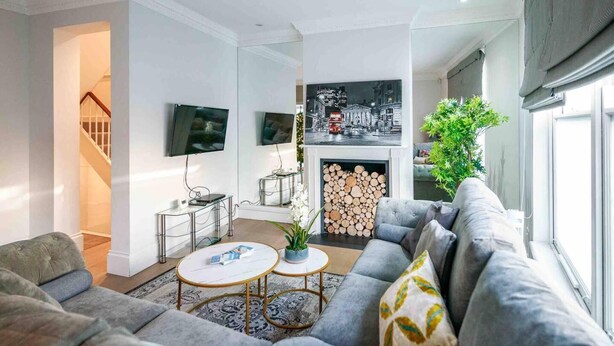 A modern living room with a grey sofa, a coffee table, a TV mounted on the wall, and a fireplace with a log fire. - Spacious & Modern 3brs House in Central London (London)