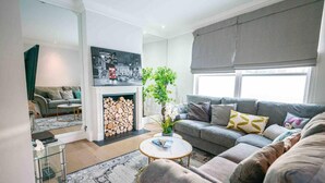 A modern living room with a grey sofa, a coffee table, a fireplace with logs, and a large window with blinds. - Spacious & Modern 3brs House in Central London (London)