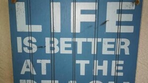 Un letrero azul con texto blanco que dice 'LIFE IS BETTER AT THE BEACH'.