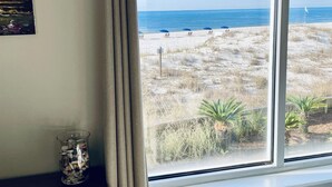 Una habitación con un amplio ventanal que da a una playa, una pequeña mesa con un recipiente de vidrio y un cuadro en la pared.
