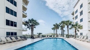 Una piscina con sillones de descanso y palmeras, rodeada por un edificio blanco con balcones.