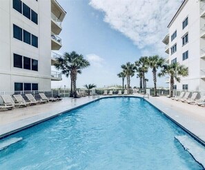 A swimming pool with lounge chairs and palm trees, surrounded by a white building with balconies.
