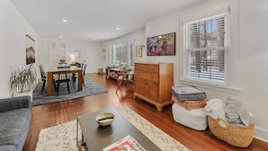 A modern living room with a dining area, wooden furniture, and a large window with blinds.