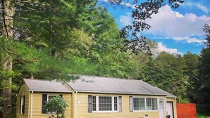 A single-story yellow house with a gabled roof, a front porch, and a small front yard with mulch and shrubs.