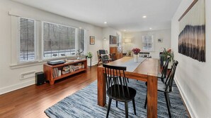 A spacious dining area with a wooden table and chairs, a bookshelf, and a large window with blinds.