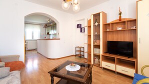 A living room with a glass-top coffee table, wooden shelving, a flat-screen TV, and a kitchen in the background. - 2-bedroom Krk Apartment - Near Krk Beaches & Town! (Krk)