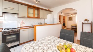 A modern kitchen with a gas stove, oven, and white cabinets. There is a dining table with a lace tablecloth and a bowl of fruit. - 2-bedroom Krk Apartment - Near Krk Beaches & Town! (Krk)