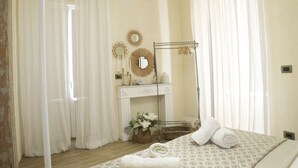 Ein Hotelzimmer mit Bett, Schminktisch mit Spiegel, kleinem Tisch und Fenster mit transparenten Vorhängen.