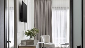 Una habitación de hotel moderna con un ventanal grande, un televisor de pantalla plana montado en la pared, una silla blanca, una mesita redonda pequeña y una planta decorativa sobre una mesita de noche de madera.