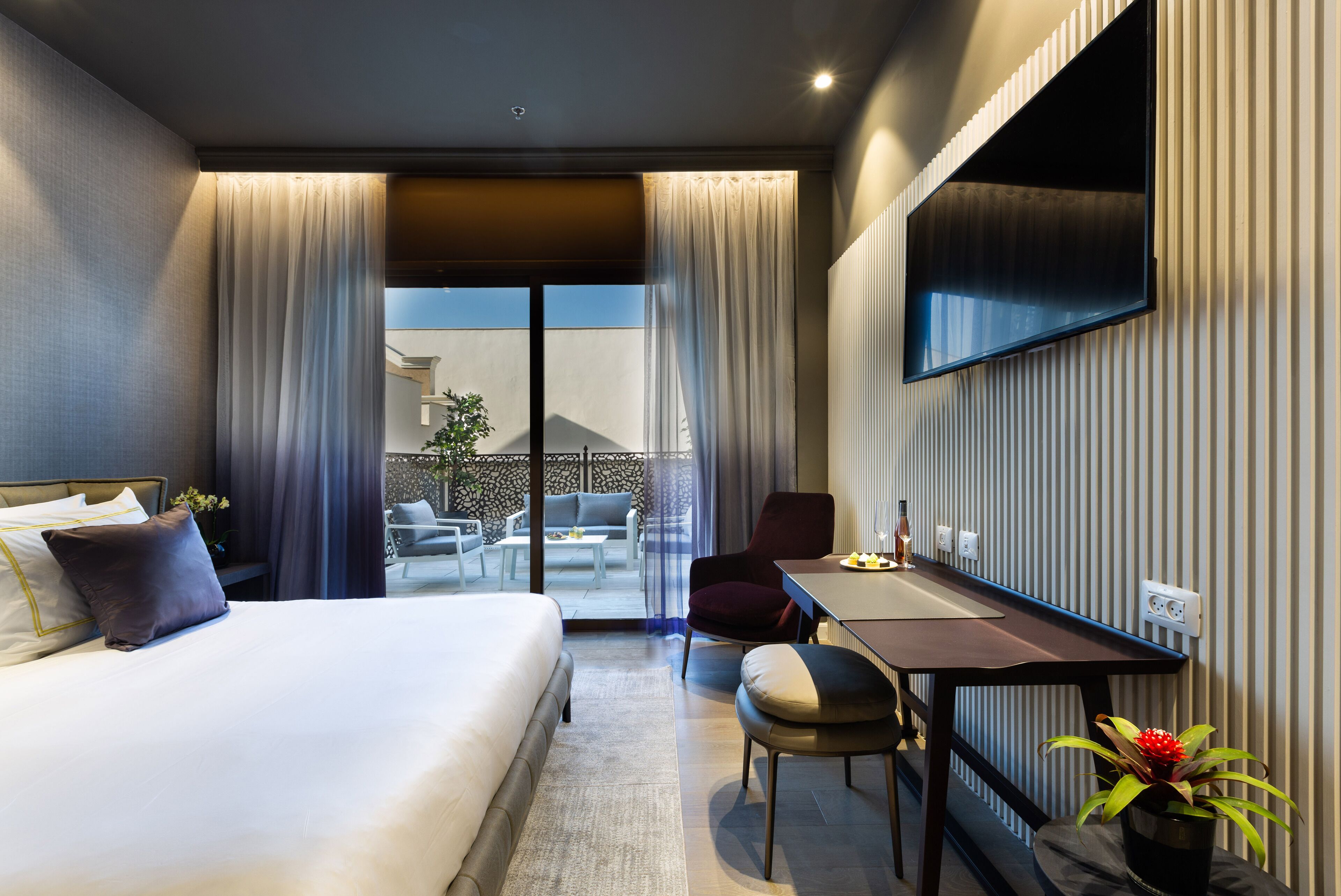 A modern hotel room with a large bed, a desk, a chair, a TV mounted on the wall, and a balcony with outdoor furniture.