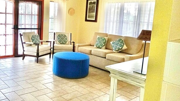 Sala de estar com sofá, cadeiras, um banquinho azul, uma mesa e uma porta que dá para o exterior.